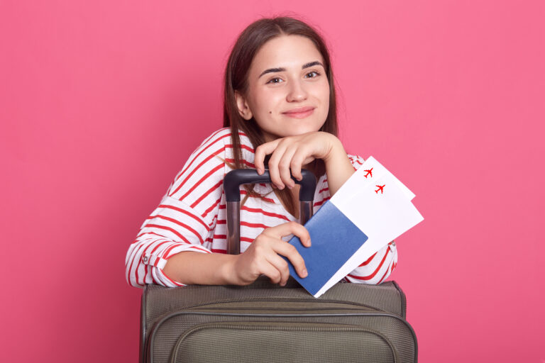 Happy woman girl with suitcase and passport isolated over pink background, dark haired girl in striped shirt looks at camera, wearing striped casual t shirt, being ready to travel.