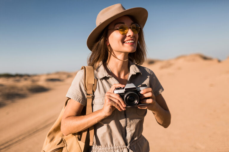 stylish young happy smiling woman in khaki dress walking in desert traveling in Africa on safari wearing hat and backpack taking photo on vintage camera, exploring nature, hot summer day, sunny weather, traveler on vacation